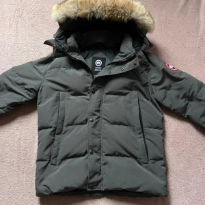 Grå pufferjacka från Canada Goose - Säljer en grå pufferjacka från Canada Goose med stor huva och snygg pälsdetalj. Jackan har två stora fickor framtill, dragkedja och knappar. Klassisk logga på ärmen och quiltad design som håller dig varm under vintern.