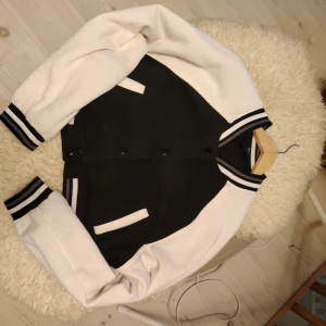 Svartvit Collegejacka från H&M barnavdelning - Snygg svartvit bomberjacka från H&M med croppad passform. Jackan har vita ärmar, svart kropp och coola randiga muddar vid hals, ärmslut och nederkant. Stängs med svarta tryckknappar och har två vita fickor framtill. Perfekt för en streetstyle-look.