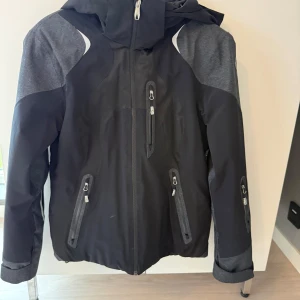 Svart och grå skidjacka från Spyder - Snygg svart och grå skidjacka från Spyder med huva och flera praktiska dragkedjefickor. Jackan har sportig design med vita detaljer och långärmade ärmar. Perfekt för dig som gillar att vara aktiv utomhus och vill ha en cool look. Snölås och en riktigt snygg skidjacka! 
