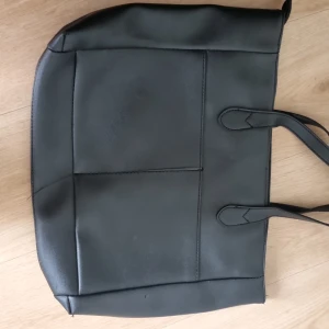 Svart stilren axelväska i skinn - Snygg svart axelväska i skinnimitation med minimalistisk design och raka linjer. Väskan har två smala handtag och en praktisk dragkedja upptill. Perfekt storlek för laptop, böcker eller andra essentials. Enkel att matcha med olika outfits.