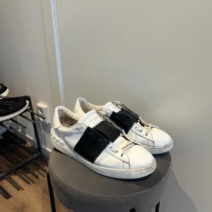 Valentino Open  - Snygga Valentino Open sneakers i vitt skinn med bred svart läderdetalj över vristen. Klassisk rund tå, platt sula och coola nitar baktill på hälen. Perfekta för dig som gillar exklusiva och stilrena sneakers med en twist. Skorna är i fint skick. Medföljer box och även orginal snören. Storlek: 45. Har även äkthetsbevis! Mvh