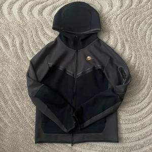 Svart vindjacka från Nike med huva - Nike vindjacka i svart med mörkgrå detaljer och gul logga på bröstet. Jackan har hel dragkedja, huva och långärmade ärmar. Materialet är syntet och är i bra skick! 