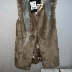 Beige och grå pälsväst från alexandria - Säljer en fluffig pälsväst från alexandria i beige och grått med snygga kontrasterande ränder. Västen är gjord i mjuk fuskpäls och har ett glansigt, champagnefärgat innerfoder. Perfekt för att ge din outfit extra edge och värme.storlek 34
