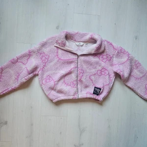 Rosa teddyjacka Hello Kitty H&M - Supergullig rosa teddyjacka från H&M x Hello Kitty med broderad Hello Kitty-patch framtill. Jackan är croppad, har dragkedja och är gjord i mjuk fleece med Hello Kitty-mönster i rosa nyanser. Perfekt för dig som vill ha en mysig och söt look.