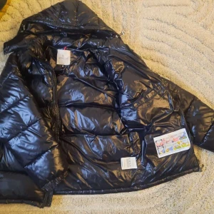 Svart Moncler pufferjacka med huva - Säljer en svart pufferjacka från Moncler med glansig finish och huva. Jackan har dragkedja framtill, quiltad design och klassisk Moncler-logga på ärmen. Perfekt för kalla dagar och riktigt snygg streetstil.