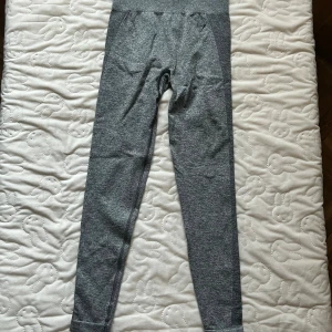 Grå leggings från Gymshark - Snygga grå leggings från Gymshark med hög midja och diskret logga på höften. De är tillverkade i ett stretchigt och mjukt material som sitter tight och bekvämt. Perfekta för träning eller chill. Melerad grå färg med mörkare paneler längs sidorna för extra stil.