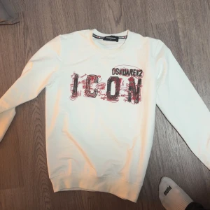 Vit Dsquared2 ICON sweatshirt - Vit sweatshirt från Dsquared2 med stort 'ICON'-tryck i svart och rött framtill. Tröjan har rund halsringning, långa ärmar och ribbade muddar. Perfekt statement-plagg för dig som gillar streetstyle och vill sticka ut.