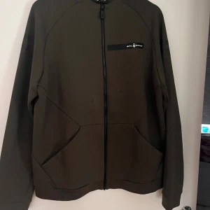 Olivgrön ziptröja från Sail Racing - Olivgrön långärmad ziptröja från Sail Racing med svart logga på bröstet. Tröjan har hel dragkedja framtill och två stora fickor. Tillverkad i en mix av polyester och bomull, vilket gör den både mjuk och slitstark. Perfekt för lager-på-lager under kyliga dagar.