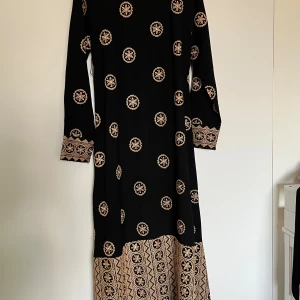 Svart långklänning med broderier - Svart långklänning med detaljerade guldbeiga broderier och mönster över hela klänningen. Långärmad modell med markerad midja och vackra broderier vid ärmslut, halsringning och nederkant. Klänningen har en traditionell och unik look.