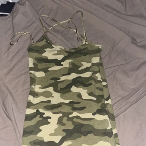 Camo kortklänning från FB Sister - Säljer en camouflagemönstrad kortklänning från FB Sister i storlek XXS. Klänningen har tunna axelband och ärmlös design, perfekt för en cool och avslappnad stil. Färgerna går i grönt, beige och svart. Materialet är mjukt och stretchigt, troligen bomull eller polyester.