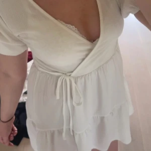 Vit omlottklänning från Gina Tricot - Supersöt vit omlottklänning från Gina Tricot med kort ärm, knyt i midjan och volangdetaljer. Klänningen har v-ringning och är tillverkad i ett lätt tyg som känns luftigt. Perfekt för varma dagar och somriga vibes!