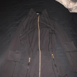 Svart träningsjacka från Lululemon - Svart figursydd träningsjacka från Lululemon med guldfärgade dragkedjor och markerade sömmar. Jackan har hög krage, långa ärmar och praktiska fickor med dragkedja. Perfekt för dig som vill ha en sportig och stilren look.