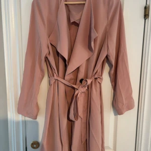 Rosa trenchcoat från Lindex, stl M - Säljer en snygg rosa trenchcoat från Lindex i storlek M. Jackan har långa ärmar, bred krage och knytskärp i midjan för en chill och trendig look. Perfekt för vår och höst när du vill ha en lätt och stilren jacka.
