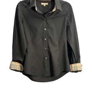 Svart Burberry skjorta  - Svart skjorta från Burberry med klassiska rutiga detaljer på insidan av krage och manschetter. Skjortan har knappar framtill, lång ärm och är tillverkad i bomull med lite stretch. Snygg och tidlös modell med en loose passform. Storlek S