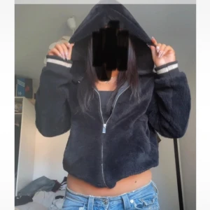 Svart teddyjacka från FB Sister - Mysig svart teddyjacka från FB Sister med huva och dragkedja framtill. Jackan är croppad och har ribbade muddar med vita detaljer vid ärmslut och nederkant. Perfekt för dig som gillar en chill och trendig stil.