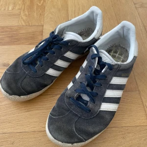 Adidas Gazelle mörkblå sneakers 37⅓ - Säljer ett par klassiska Adidas Gazelle sneakers i mörkblå mocka med vita detaljer och de ikoniska tre ränderna på sidorna. Skorna har blå snörning, vit plös och vit insida. Perfekta för dig som gillar retrostil och streetwear. Saknar innersula