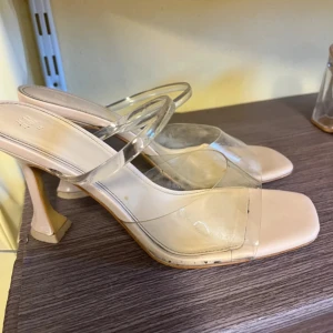 Beige klackpumps med transparent rem - Säljer ett par stilrena pumps från H&M i beige med transparenta plastremmar. Skorna har fyrkantig tå och hög klack med en modern form. Perfekt för dig som gillar minimalistisk och trendig design. Materialet är syntet och plast. Finns defekter se bilder.