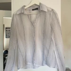 Vit randig skjorta från Mexx - Snygg vit skjorta från Mexx med tunna svarta ränder och diskreta prickar. något genomskinlig. Skjortan har klassisk krage, knappar framtill och är långärmad. Materialet är lätt och luftigt, perfekt för varmare dagar. Passformen är normal och skjortan har en stilren look.