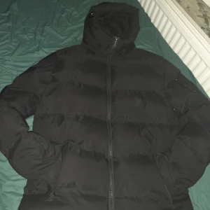 Svart pufferjacka Supply & Demand  - Svart pufferjacka från Supply & Demand med luva och dragkedja. Jackan har en clean look med logga på ärmen och text på luvan. Perfekt för kalla dagar och riktigt skön passform. Fodrad insida och praktiska fickor.