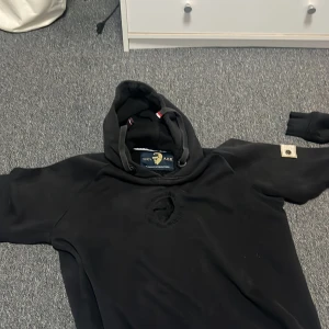 Svart hoodie från WHO'S A.C.E - Svart hoodie från WHO'S A.C.E med broderad logga på bröstet och snygg patch med knappar på ärmen. Tröjan har huva med snörning och coola detaljer i guld. Perfekt för dig som gillar streetwear och vill sticka ut.