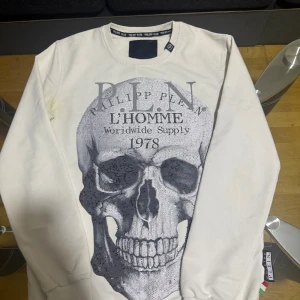 Vit sweatshirt med döskalle från Philipp Plein - Säljer en vit sweatshirt från Philipp Plein med stor grå döskalle och text framtill. Tröjan har rund hals, långa ärmar och ribbade muddar. Snygg detalj med metallogo i nacken och etikett med italienska flaggan vid nederkanten.