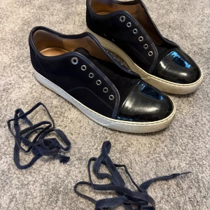 Lanvin svarta sneakers med lackad tå - Snygga svarta sneakers från Lanvin med vit sula och lackad tå. Ovandelen är i mörk mocka och insidan är i beige skinn. Skorna har rund tå och medföljande svarta snören. Perfekt för dig som gillar stilrena och lyxiga sneakers.