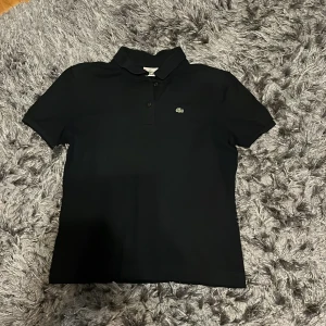 Svart pikétröja från Lacoste - Klassisk svart pikétröja från Lacoste med korta ärmar. Tröjan har krage och knäppning framtill. Perfekt för en clean och sportig look.