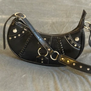 Svart axelväska i skinn med nitar - Cool svart axelväska i skinn med silvriga nitar, ringdetaljer och dekorativa sömmar. Väskan har en böjd form, justerbart axelband och dragkedja upptill. Perfekt för dig som gillar edgy och unik stil. Dragkedjan funkar👍🏼