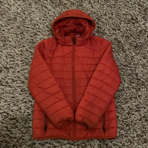 Röd dunjacka från Everest - Säljer en röd dunjacka från Everest i storlek 158/164. Jackan har huva, dragkedja framtill och två sidofickor med dragkedja. Quiltad design och tillverkad i syntetmaterial, perfekt för kyliga dagar.