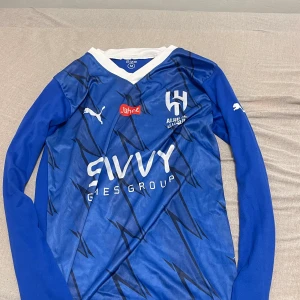 Blå Al Hilal Neymar Jr fotbollströja M - Säljer en blå långärmad Al Hilal fotbollströja med Neymar Jr och nummer 10 på ryggen. Tröjan har vita detaljer, V-ringad krage och tryck från Puma, Jahez och Savvy. Materialet är lätt och andas, perfekt för match eller träning. Snyggt grafiskt mönster framtill.