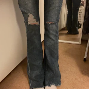 Mörkblå bootcut jeans med slitningar - Säljer ett par mörkblå bootcut jeans med slitna detaljer på knäna och råa kanter nertill. Jeansen har en klassisk femficksmodell och är tillverkade i denim. Perfekta för dig som gillar en avslappnad och trendig look.