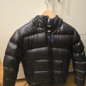 Svart pufferjacka med huva - Säljer en svart pufferjacka från Everest med blått foder och huva. Jackan har dragkedja framtill och två fickor med dragkedja. Perfekt för kalla dagar och riktigt skön att ha på sig.