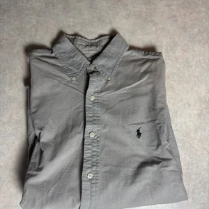 Grå skjorta Polo Ralph Lauren M - Klassisk grå skjorta från Polo Ralph Lauren i slim fit-modell. Skjortan har knappar framtill, lång ärm och den ikoniska broderade loggan på bröstet. Tillverkad i mjuk bomull och passar perfekt för en clean och stilren look.
