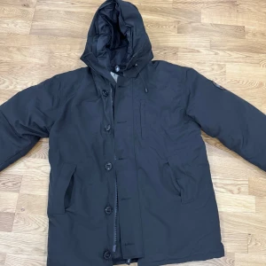 Svart dunjacka från Canada Goose - Säljer en svart dunjacka från Canada Goose i storlek L. Jackan har huva, dragkedja och knappar framtill samt två fickor på sidorna. Perfekt för kalla dagar och har en klassisk, stilren look med diskret logga på ärmen.