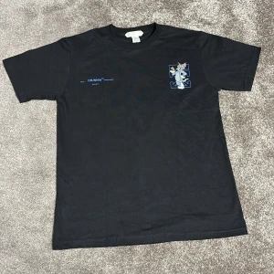 Off-White x Tom & Jerry svart t-shirt - Svart t-shirt från Off-White med Tom & Jerry-tryck på ryggen och blå logga. Klassisk passform, kortärmad och tillverkad i 100% bomull. Snygg detalj med stor blå Off-White-pil och tecknad grafik. Perfekt statement-plagg för dig som gillar streetwear.