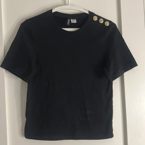 Svart topp med guldknappar från H&M - Svart kortärmad topp från H&M Divided med rund hals och tre snygga guldknappar på ena axeln. Toppens material är mjuk bomull och modellen är croppad med en normal passform. Perfekt för dig som gillar stilrena detaljer.
