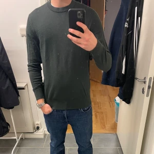 Mörkgrön stickad tröja från H&M - En stilren mörkgrön stickad tröja från H&M med rund halsringning och långa ärmar. Tröjan har en klassisk passform och ribbade muddar vid ärmslut och nederkant. Perfekt för lager-på-lager och enkel att matcha med jeans eller chinos. Skick 9/10