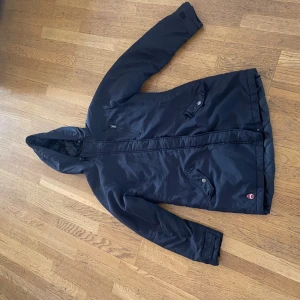 Svart parkajacka från Esprit med huva - Svart parkajacka från Esprit med justerbar huva, dragkedja och tryckknappar. Jackan har två stora fickor framtill och är vadderad för extra värme. Perfekt för kyliga dagar och har en stilren, enkel look.