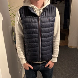 Mörkblå dunväst från Tommy Hilfiger - Snygg mörkblå dunväst från Tommy Hilfiger med quiltad design och dragkedja framtill. Västen har diskreta logodetaljer och blått foder på insidan. Perfekt att slänga över en hoodie för en chill look.