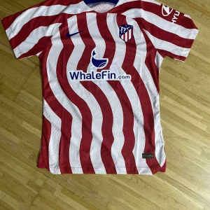 Atlético Madrid matchtröja Nike röd/vit - Säljer en officiell Atlético Madrid fotbollströja från Nike med vågiga röda och vita ränder, klubbmärke på bröstet och sponsortryck. Tröjan är kortärmad och tillverkad i lätt, ventilerande polyester – perfekt för match eller träning.