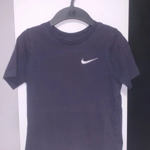 Mörkblå Nike t-shirt med logga - En stilren mörkblå t-shirt från Nike med klassisk vit swoosh-logga på bröstet. T-shirten har rund halsringning och korta ärmar, perfekt för en sportig och avslappnad look.