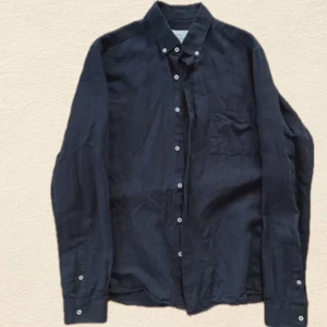 Marinblå linneskjorta med knappar - Stilren svart skjorta i linne med klassisk krage och vita knappar framtill. Skjortan har lång ärm och en bröstficka, perfekt för en clean och avslappnad look.