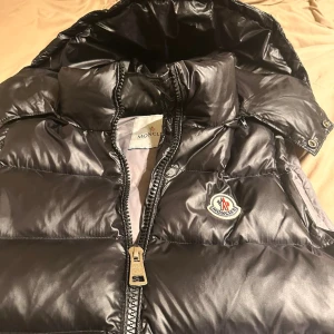 Svart dunväst från Moncler med huva - Snygg svart dunväst från Moncler med glansig finish, avtagbar huva och dragkedja framtill. Västen har två fickor med dragkedja och klassisk Moncler-logga på bröstet. Perfekt för lager-på-lager och streetstyle.
