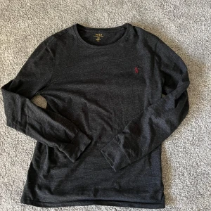Mörkgrå långärmad tröja Polo Ralph Lauren - Snygg mörkgrå långärmad tröja från Polo Ralph Lauren med klassisk rund hals och röd broderad logga på bröstet. Tröjan har en normal passform och är tillverkad i mjuk bomull, perfekt för lager-på-lager under höst och vinter.