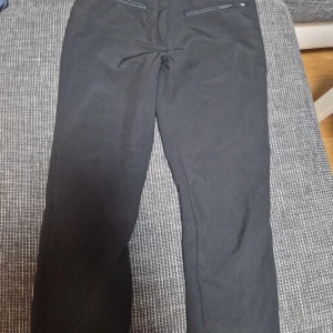 Svarta täckbyxor från Icepeak, strl 36 - Svarta täckbyxor från Icepeak med dragkedja och knäppning fram, fleecefoder på insidan och praktiska resårband vid benslut. Perfekta för kalla dagar och vinteraktiviteter. Snygg och enkel design med raka ben och diskreta fickor med dragkedja.