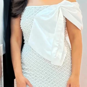 Vit långklänning med stor rosett - Supersnygg vit långklänning med offshoulder och en stor rosett framtill. Klänningen har ett glittrigt rutmönster över hela tyget och sitter tight för en elegant siluett. Perfekt för dig som vill sticka ut med en unik detalj och glamorös vibe. Nypris: 2600kr (med tull) använd endast en gång! S men kan också passa en liten M. 