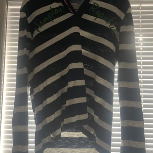 Randig långärmad tröja Hampton Republic - Säljer en långärmad tröja från Hampton Republic med breda marinblå och vita ränder. Tröjan har grönt tryck på bröstet, knappar vid halsen och klassisk krage med rödvit detalj. Perfekt för en chill och stilren look.