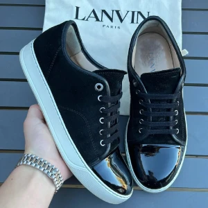 Svarta sneakers från Lanvin med lackad tå - Snygga svarta sneakers från Lanvin med vit sula och glansig lackad tå. Ovandelen är i mocka och har klassisk snörning med metallöglor. Skorna har en rund tå och en stilren design som sticker ut med den blanka detaljen framtill.
