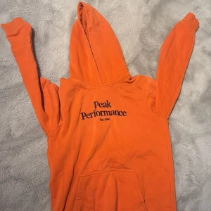Orange hoodie från Peak Performance - Säljer en orange hoodie från Peak Performance med svart logga på bröstet. Tröjan har huva, magficka och långa ärmar. Perfekt för dig som gillar sportig och avslappnad stil.