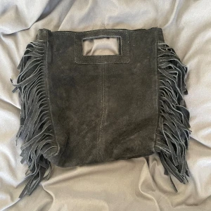 Svart mocka handväska med fransar - Cool svart handväska i mocka med fyrkantigt handtag och långa fransar längs sidorna. Väskan har en stilren och lite bohemisk vibe, perfekt för dig som vill sticka ut. Rymmer det viktigaste och är lätt att bära i handen.